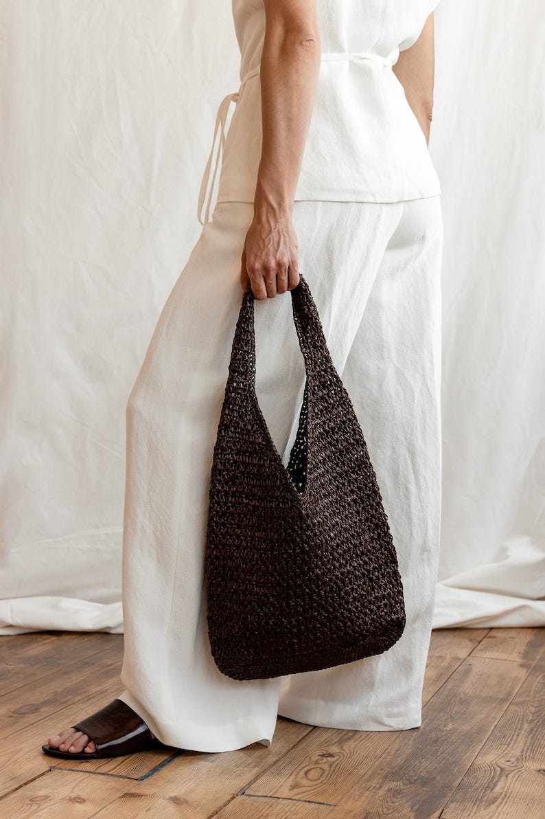May include: A dark brown woven tote bag with a wide opening is held by a person. The bag has a textured appearance and a simple, functional design. The person is wearing white pants and a white top.