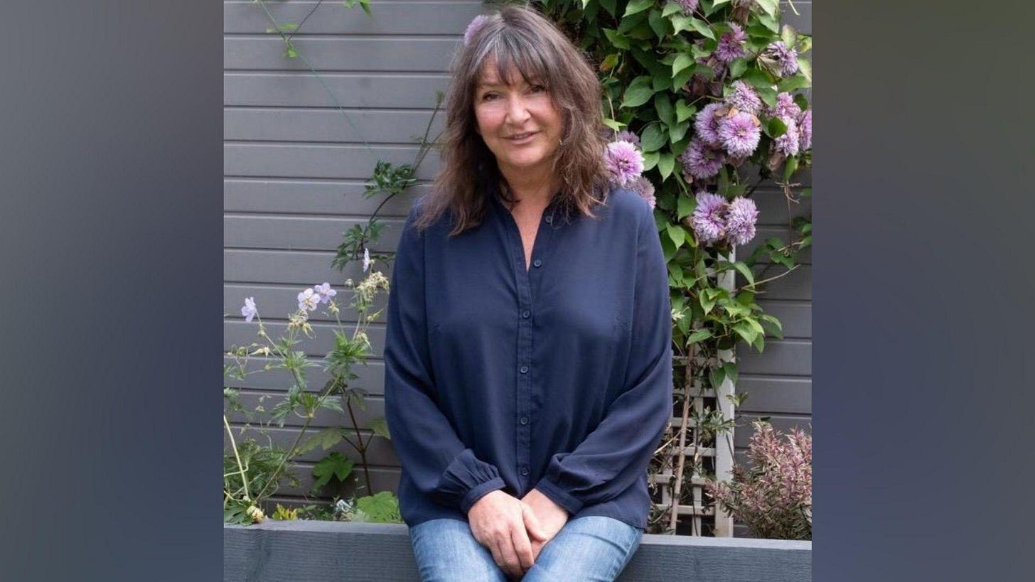 A woman in a blue shirt and jeans sits on a garden wall with flowers behind her A woman in a blue shirt and jeans sits on a garden wall with flowers behind her