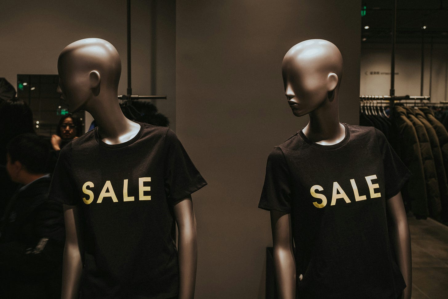 Two headless mannequins facing each other in a dimly lit retail store, both wearing black t-shirts printed with the word SALE in large gold letters, with clothing rails visible in the background.