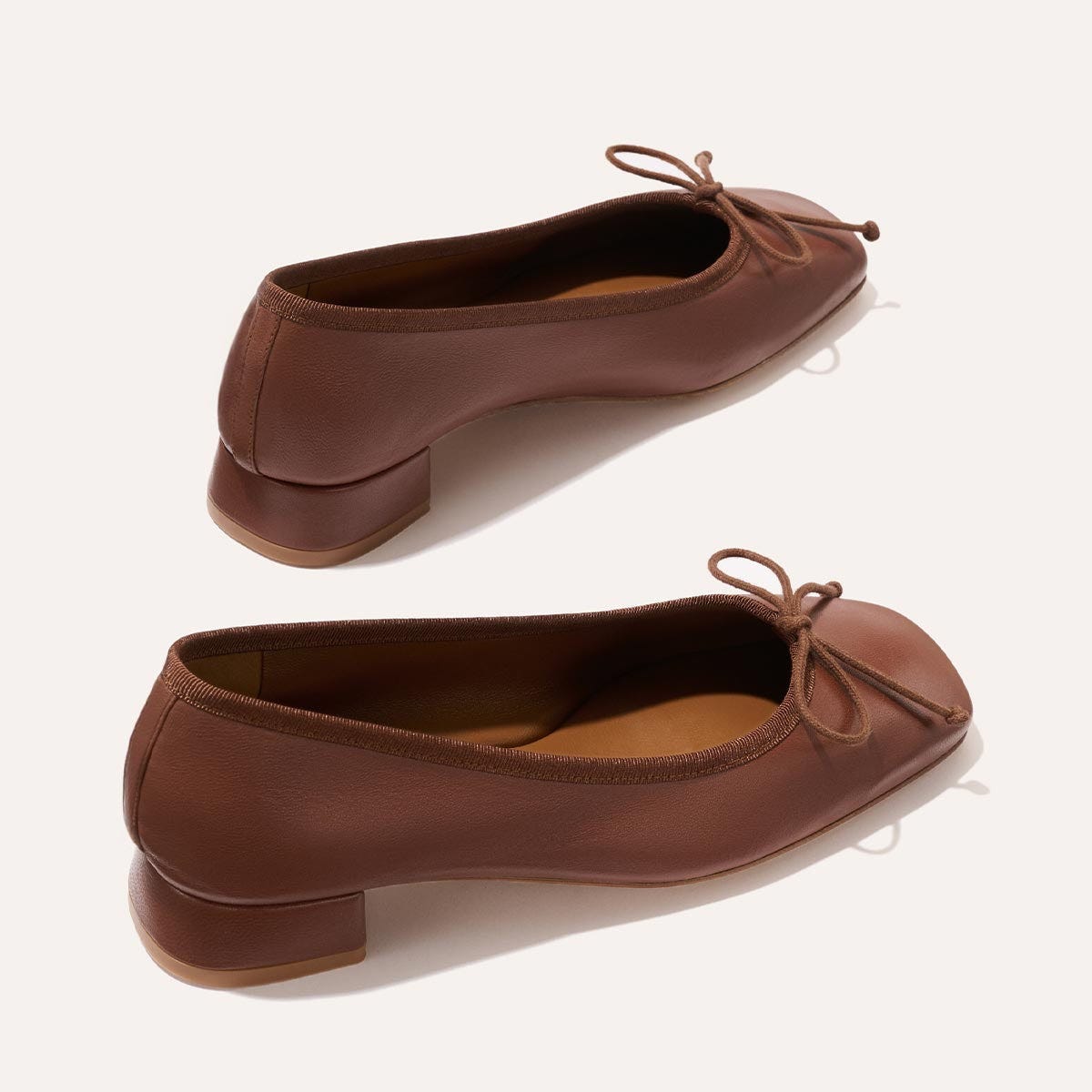 The Ada - Cognac Nappa by margaux-development: brown leather ballet pumps with low block heels, rounded toes, sacchetto construction for a glove-like fit, and thin bow ties on the front, shown on a light background. The Ada - Cognac Nappa by margaux-development: brown leather ballet pumps with low block heels, rounded toes, sacchetto construction for a glove-like fit, and thin bow ties on the front, shown on a light background.