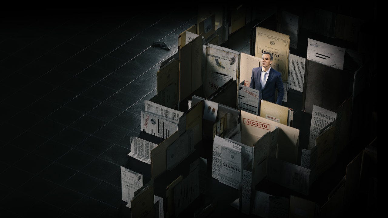 A white middle-aged man stands inside a building made of papers marked "confidential," surrounded by darkness
