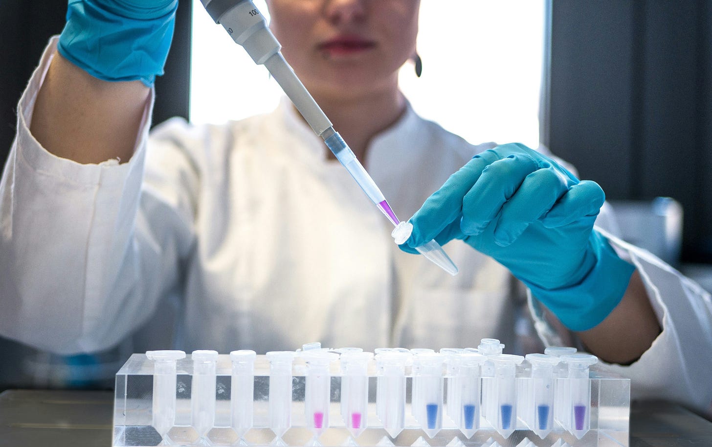 Color photo of a lab tech in a white coat behind a tray of vials, using a large syringe to drop liquid into a pipette