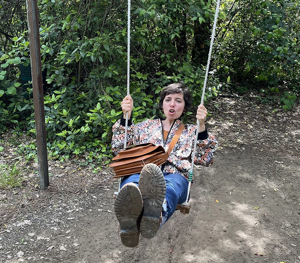 Plan moyen de ma personne sur une balançoire en extérieur, les jambes tendues vers l'avant en direction du photographe. Je porte une veste à fleurs et un sac marron en bandoulière. Le décor est un chemin de terre bordé de végétation. Plan moyen de ma personne sur une balançoire en extérieur, les jambes tendues vers l'avant en direction du photographe. Je porte une veste à fleurs et un sac marron en bandoulière. Le décor est un chemin de terre bordé de végétation.
