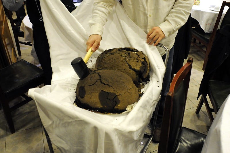 a man in chef's whites hits a clay wrapped object on top of a dining trolley and surrounded by white cloth, with a hammer a man in chef's whites hits a clay wrapped object on top of a dining trolley and surrounded by white cloth, with a hammer
