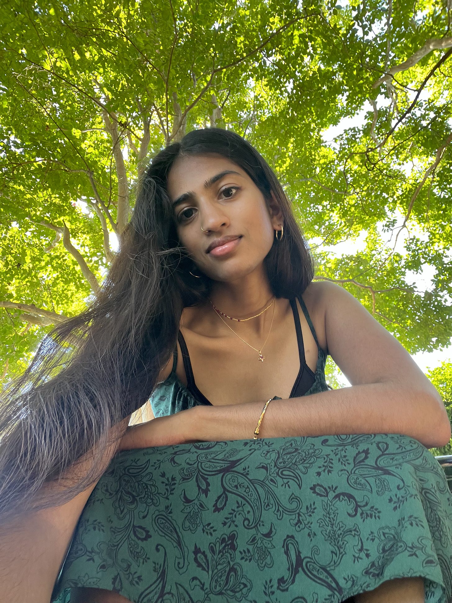 Tan, light skinned south Asian person with long black hair wearing a long forest green sundress sits underneath a bright green tree and leans into the camera which they are holding below them, slightly smiling. They are wearing a gold-toned necklace, bangle, and hoop earrings. The sun filters through the backlit trees.