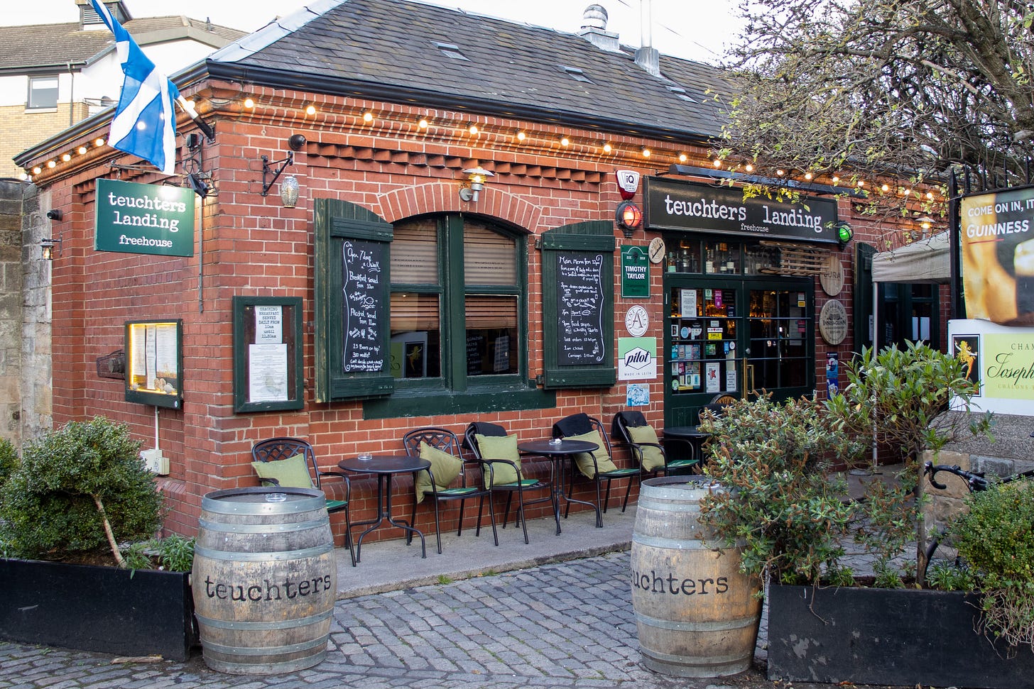 The brick exterior of a pub, with a Saltire flag hanging outside and green shutters outside the window. 