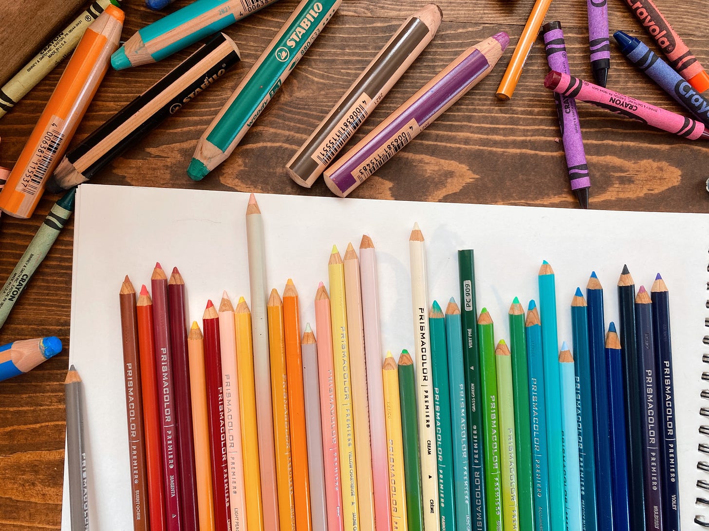 Rainbow of colored pencils on sketchbook paper surrounded by crayons and stabile pencils.