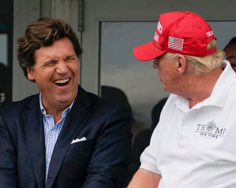 A younger man man laughs with an older man wearing a red hat with a US flag on it.