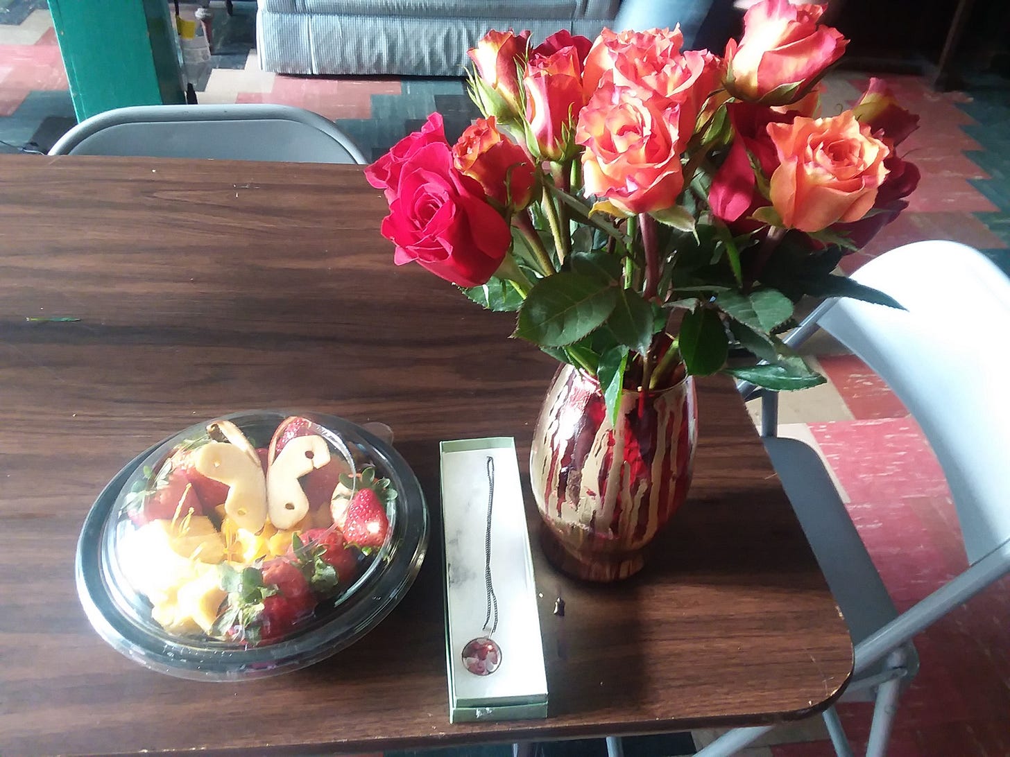 A fancy fruit bowl with apple butterflies, a necklace in a gift box, and a red and gold painted vase with red and gold roses on it sitting on an ugly brown folding table in a studio space.