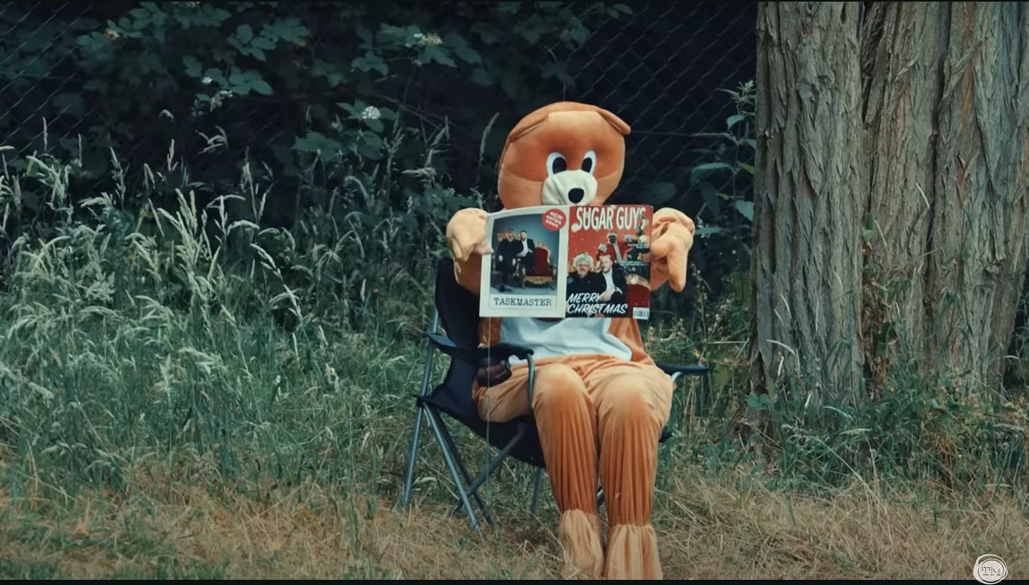 A person in a bear suit reads “Sugar Guys” magazine on a folding chair by a tree.