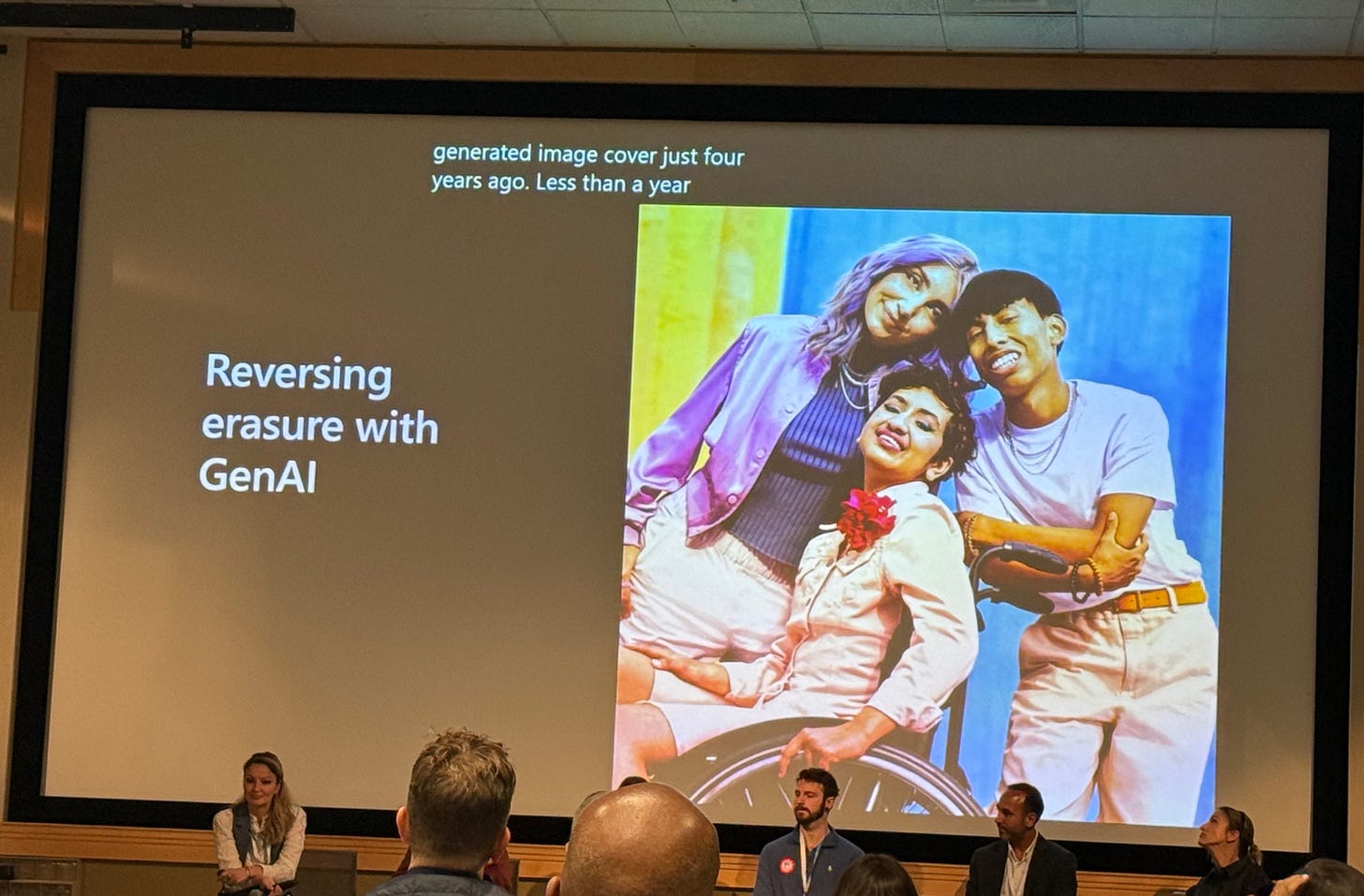 Projected slide reading "Reversing erasure with GenAI" next to a vibrant photo of three smiling young people together, one in a wheelchair, with panelists visible below. Projected slide reading "Reversing erasure with GenAI" next to a vibrant photo of three smiling young people together, one in a wheelchair, with panelists visible below.
