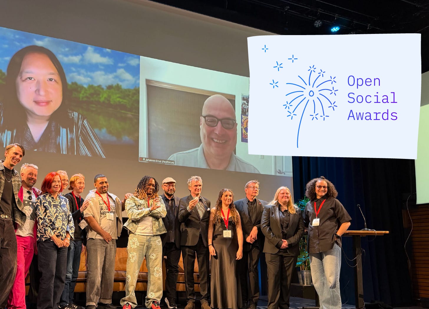 A group of about twelve people stand together on a stage, smiling. Behind them, a large projection screen shows two video call participants, one labeled "Johannes Ernst (FediForum)." To the right, a separate screen displays the Open Social Awards logo with a dandelion illustration. A podium with a microphone is visible on the right side of the stage.