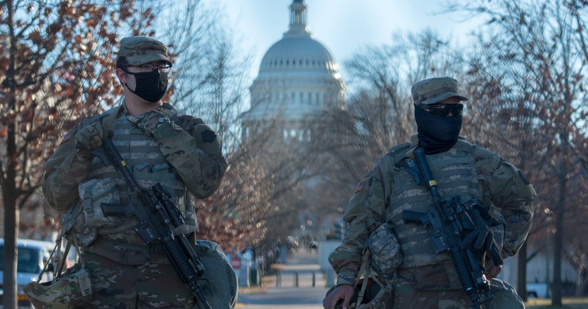 Masked National Guard Troops in full uniform carrying military assault rifles. The U.S. Capitol building is in the background.