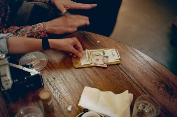 A person's hand places money on a clipboard with bills on a wooden table. Blurred hands are visible above in motion.