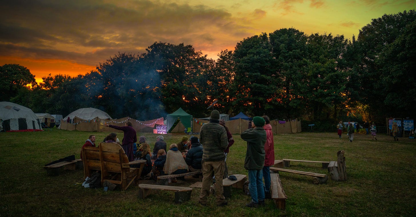 Campers gather around a fire at sunset, with tents in the background and a warm glow illuminating the sky.