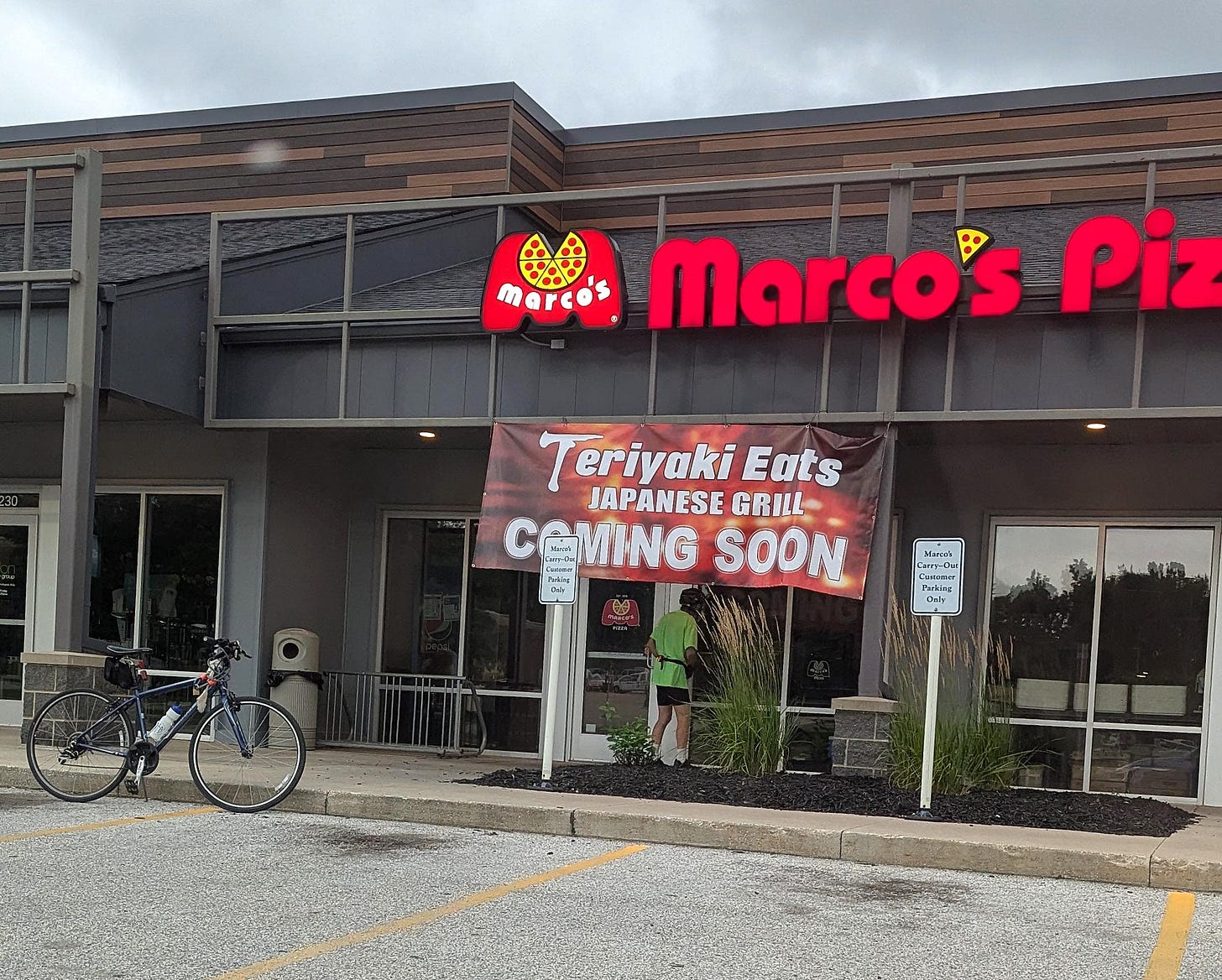 A photo of the front of a closed Marco's Pizza with a sign reading "Teriyaki Eats Japanese Grill Coming Soon" . . . and a bicyclist looking in the windows of the store