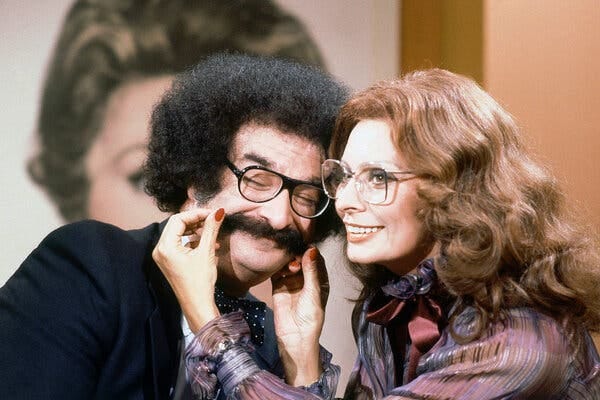 A glamorous older movie star tugs at the mustache of a man with bushy hair. A glamorous older movie star tugs at the mustache of a man with bushy hair.