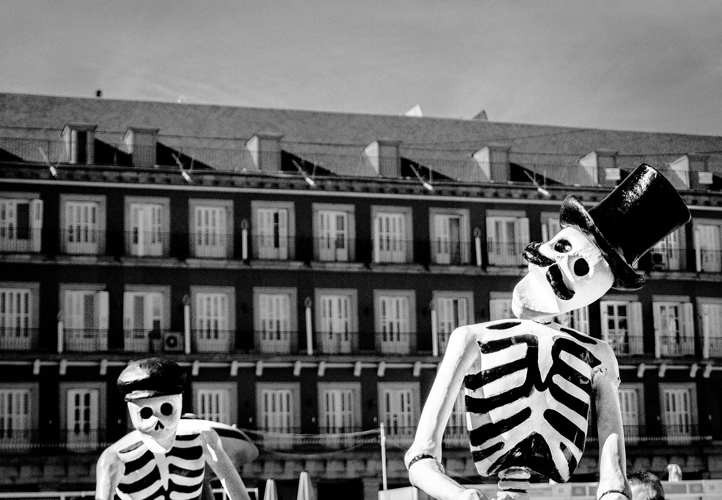 Black-and-white photograph of two skeletal figures in costumes, posed in front of a tall building with rows of identical windows. One skeleton wears a large top hat and bowtie, leaning slightly forward, while the other in the background wears a smaller hat. Both have hollow eyes and toothy grins, giving the scene a theatrical, macabre look.