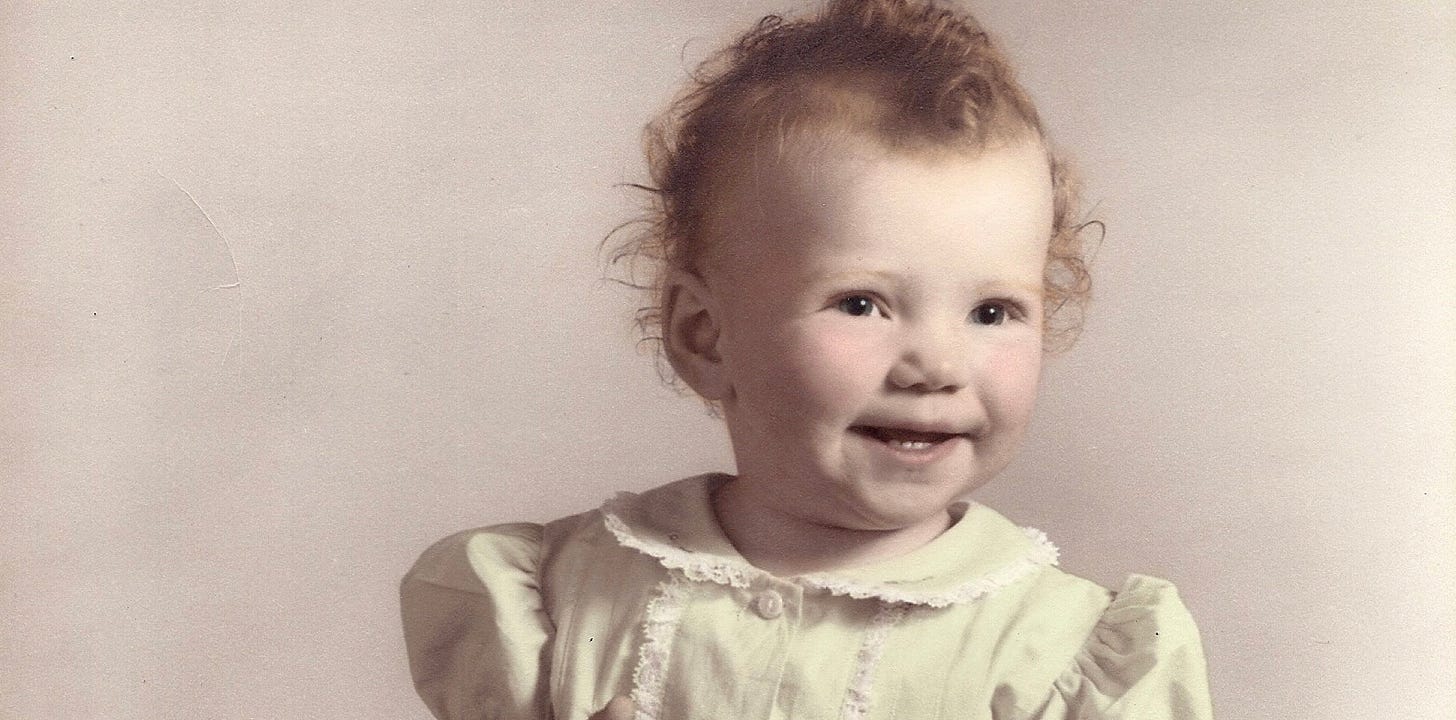 M.A. Hastings as a one-year old. She has red curly hair, blue eyes and is smiling at the camera.  She’s wearing a light lime colored dress with puffy short sleeves and a Peter Pan collar that has lace around it.