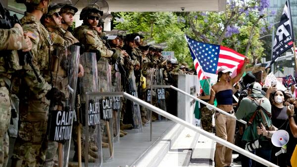 A line of National Guard troops wearing helmets and carrying shields stands on the steps of a federal building with a large crowd of protestors a few feet in front of them, One protester holds a large American flag. 