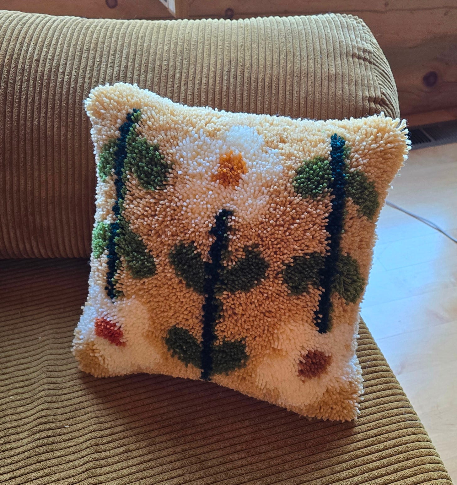 A throw pillow that is tan and green and white, with a flower pattern on it, made by latch hooking with pieces of yarn A throw pillow that is tan and green and white, with a flower pattern on it, made by latch hooking with pieces of yarn