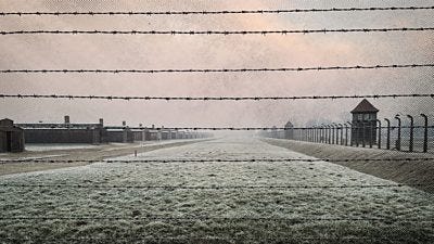 An image of a camp with a series of long buildings and huts, seen through barbed wire