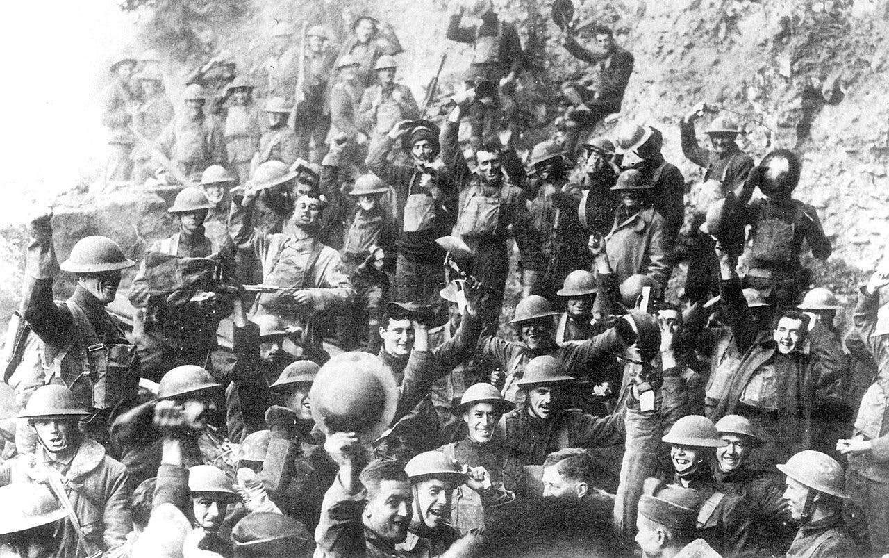 Historical black and white photograph showing jubilant World War I soldiers celebrating the news of demobilization. The troops, wearing tin Brodie helmets, are crowded together against a rocky backdrop, many with raised arms and cheerful expressions. Some are holding up bottles and waving, creating a spontaneous moment of joy captured on camera.