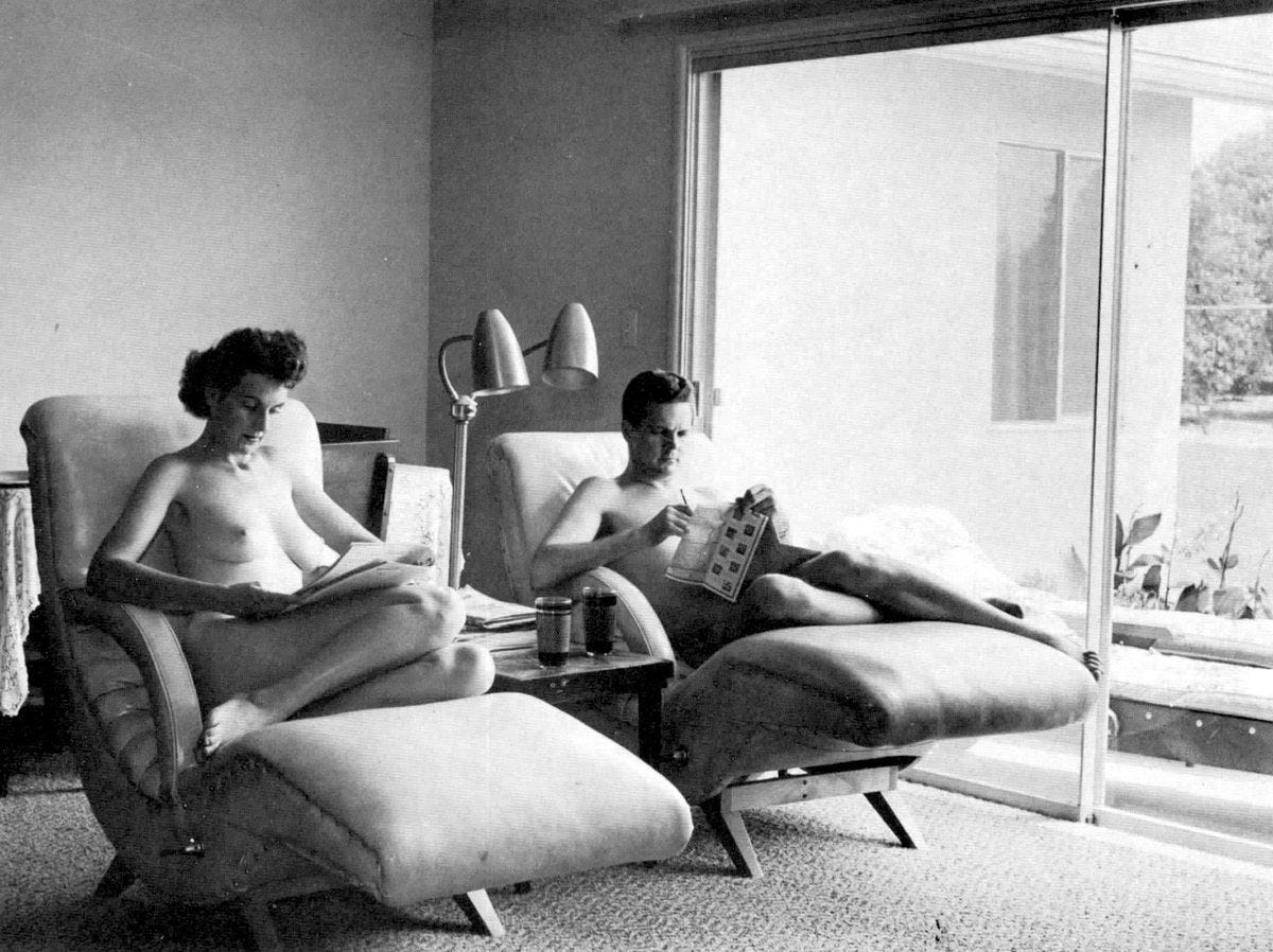 Black-and-white photograph of two nude adults reclining in cushioned lounge chairs inside a modern living room, each reading printed material. A small table with soda cans and a dual-headed floor lamp sits between them, while a large sliding glass door opens to a sunlit outdoor area, giving the scene a calm, domestic feel. Black-and-white photograph of two nude adults reclining in cushioned lounge chairs inside a modern living room, each reading printed material. A small table with soda cans and a dual-headed floor lamp sits between them, while a large sliding glass door opens to a sunlit outdoor area, giving the scene a calm, domestic feel.