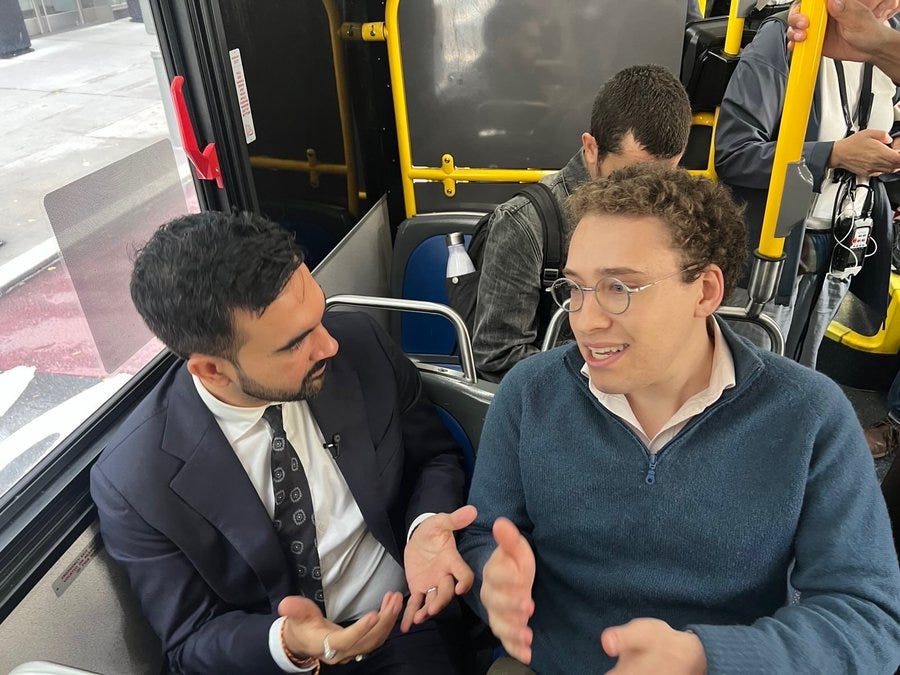 Inside a yellow and black city bus with blue seats and windows showing blurred urban streets, two men sit facing each other. The man on the left wears a dark suit, white shirt, striped tie, and has short dark hair and a beard, gesturing with both hands. The man on the right wears a light blue sweater, glasses, curly hair, and smiles while gesturing. Other passengers are visible in the background, including one with a phone and another in a gray shirt.