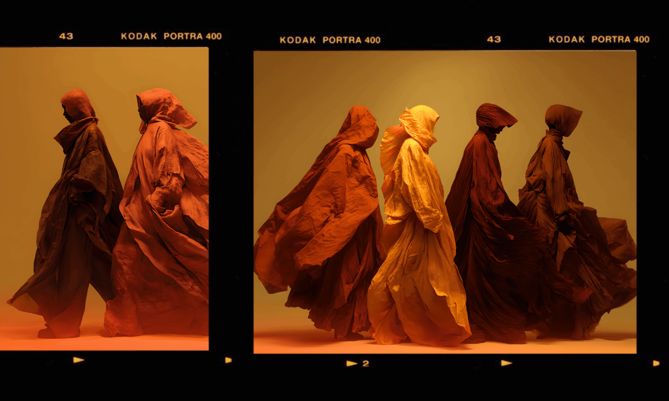 For The Subtext Review's Overtones: Four figures cloaked entirely in flowing, textured fabric (brown, orange, and yellow) stand together in diffused light, appearing to be in motion. The garments heavily obscure the bodies, emphasizing the movement of the cloth over human form.
