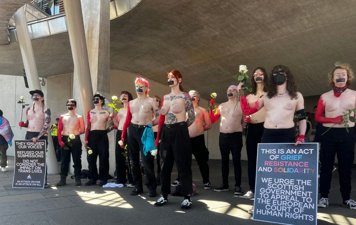At least a dozen trans and non-binary protesters stand with bare chests and black tape over their mouths. 