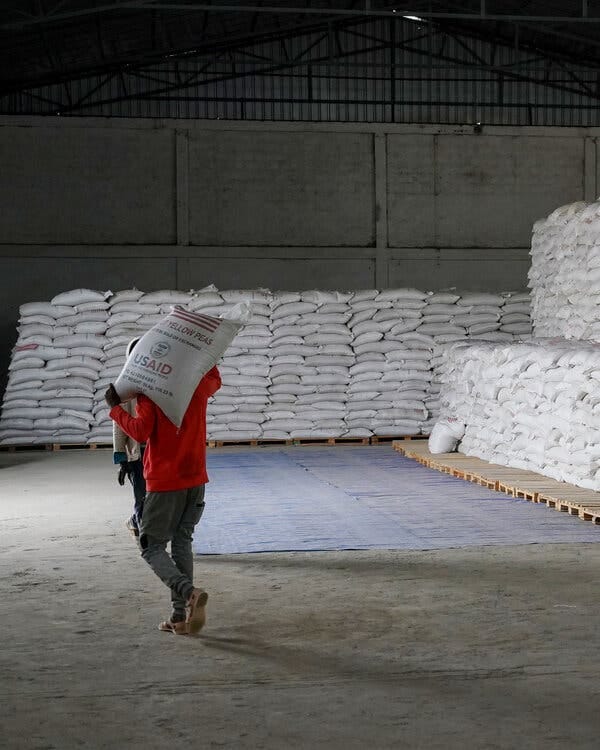 A man carries a large bag of yellow peas out of a warehouse. The bag is marked with U.S.A.I.D. A man carries a large bag of yellow peas out of a warehouse. The bag is marked with U.S.A.I.D.