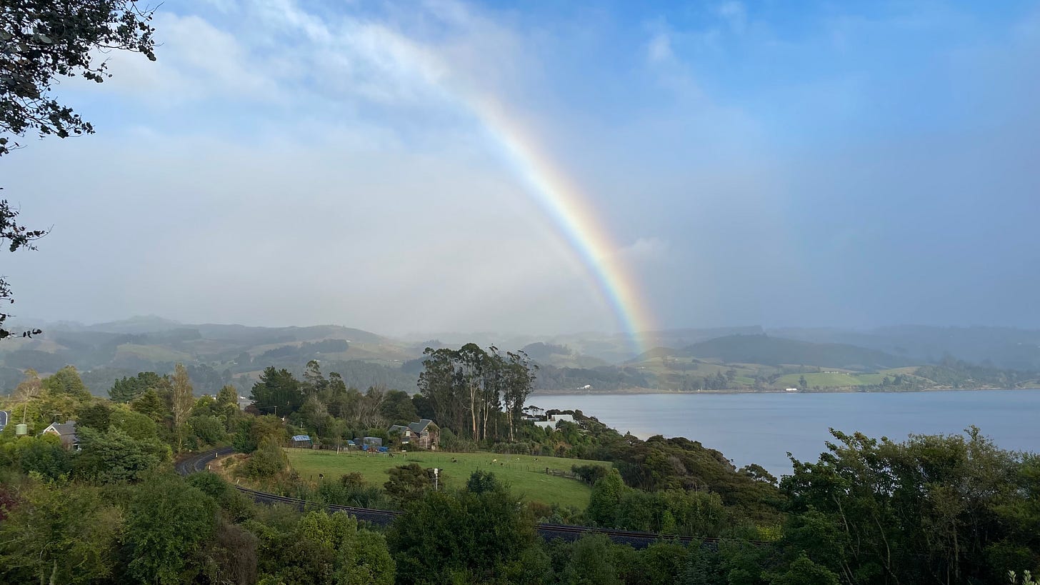 A landscape, with green fields in the foreground, water, blue sky, and a bit rainbow in the distance.