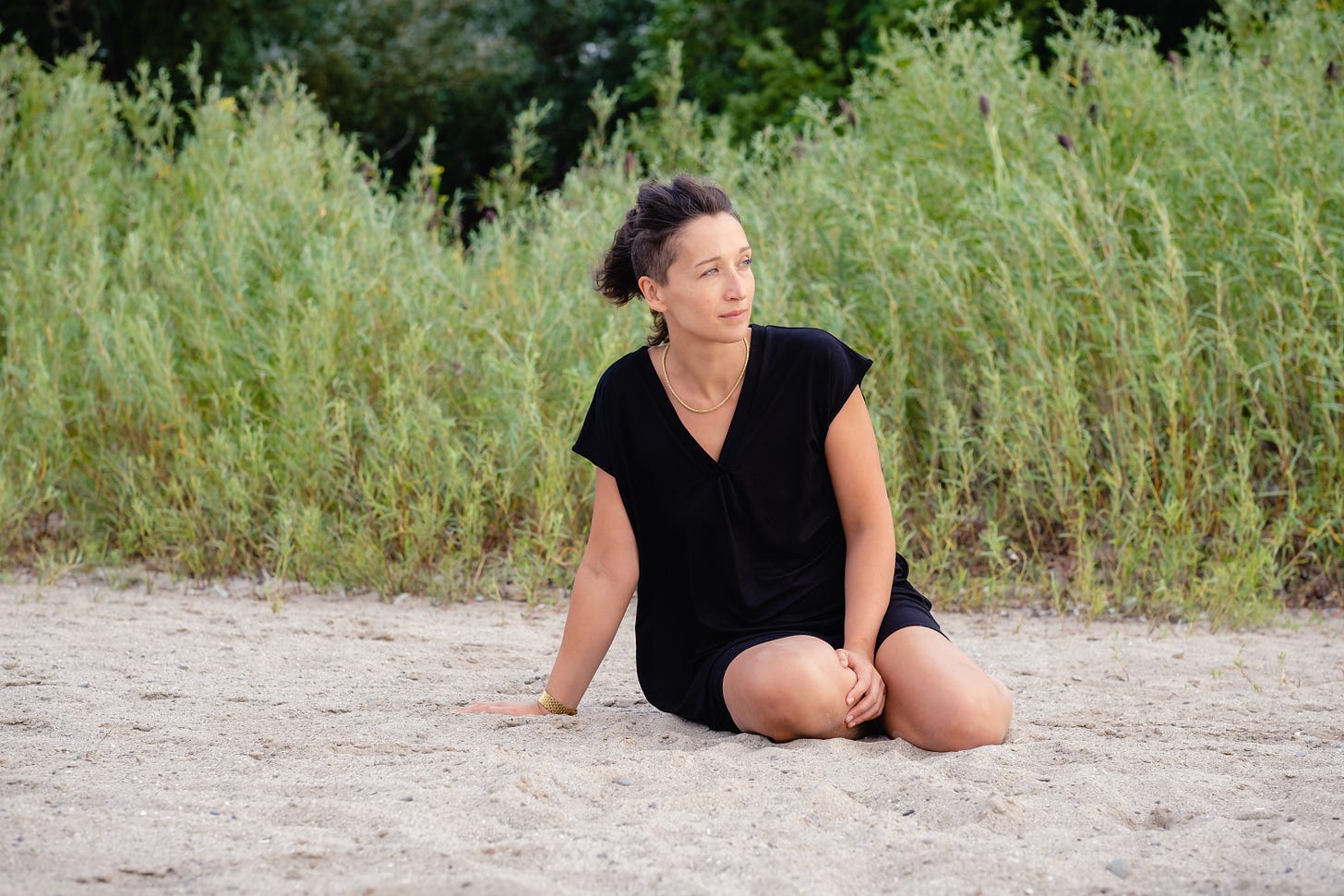 Tarzan sitting in the sand, her knees to the side, wearing a loose black v-neck dress and looking out into the distance, her hair blowing in the wind