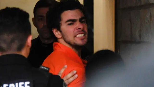 Luigi Mangione, 26, a suspect in the New York City killing of UnitedHealth executive Brian Thompson, is seen looking angry with deputies in an orange prison jumpsuit. He is at court in a hallway.  