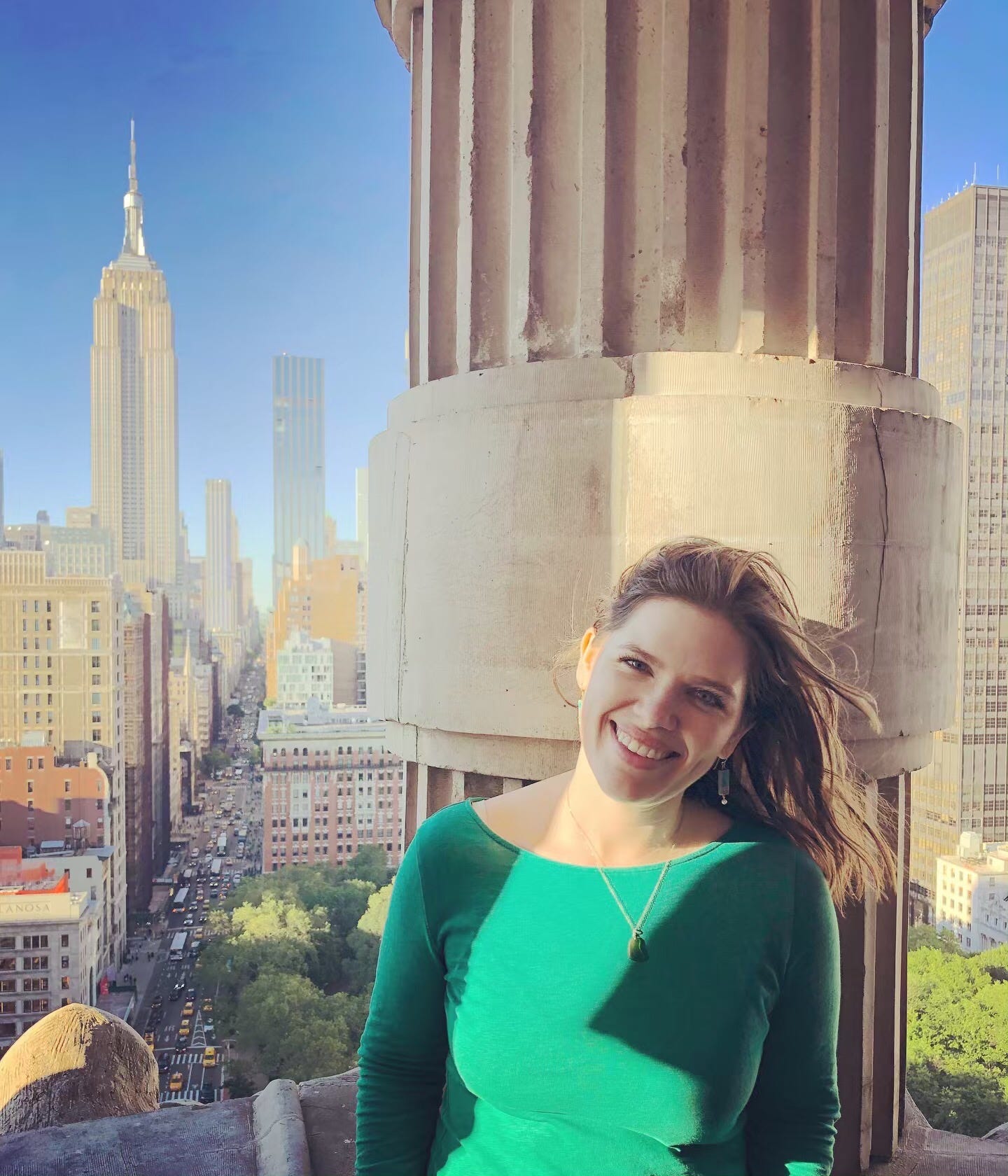 Katharine Duckett, a white woman in her thirties with long brown hair, is smiling and wearing a green long-sleeved shirt and necklace with a green stone while standing on the balcony of the Flatiron Building in Manhattan, with the Empire State Building visible in the background.
