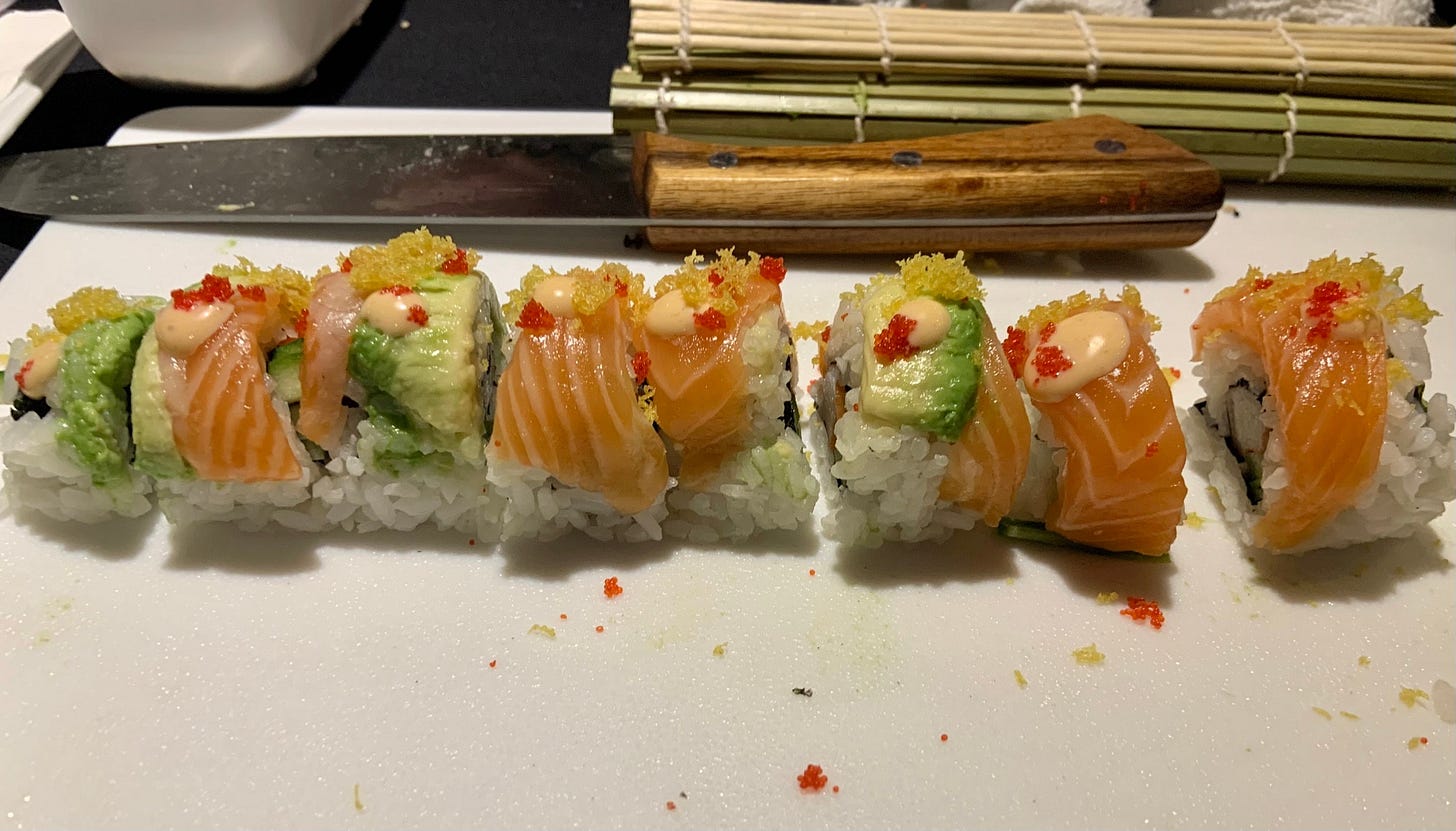 A photo of eight specialty sushi rolls on a chopping board. The rolls have avocado and salmon on the outside in an alternating striped pattern. Each one has a dot of spicy mayo, a sprinkle of tempura flakes, and red roe. The cutting board is messy, and in the background is a sharp sushi knife and a rolled-up makisu. A photo of eight specialty sushi rolls on a chopping board. The rolls have avocado and salmon on the outside in an alternating striped pattern. Each one has a dot of spicy mayo, a sprinkle of tempura flakes, and red roe. The cutting board is messy, and in the background is a sharp sushi knife and a rolled-up makisu.