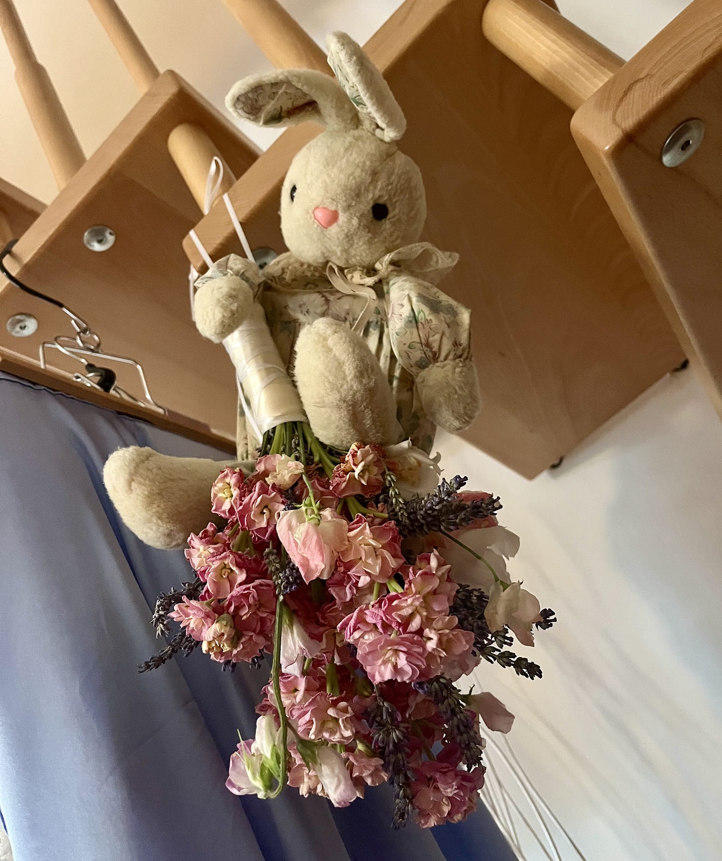Ein Brautstrauß hängt mit den Blüten nach unten an einer Treppe, in Kuschelhase reitet darauf, in Hintergrund ein hellblauer Rock.