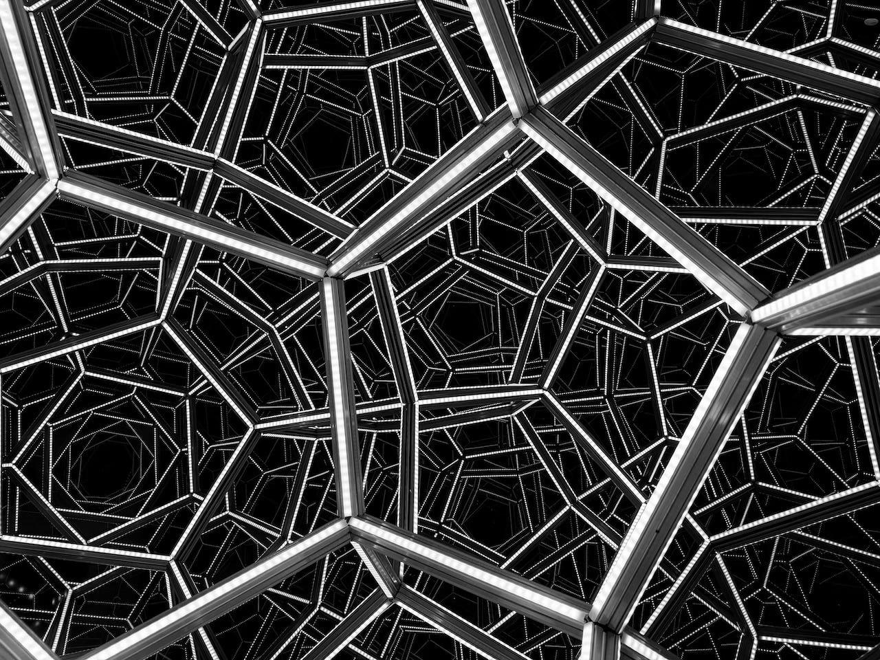 A photo of an intricate geometric lattice of metallic pentagon-shaped frames repeating into deep space against a black background, creating a tunnel-like, abstract pattern.