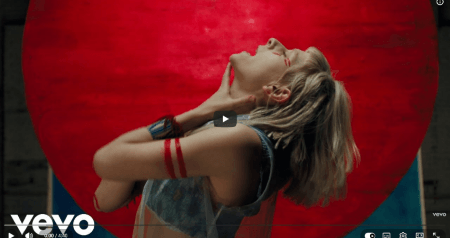 Screenshot of a music video showing a young woman with short blond hair and red war paint singing against a red background.