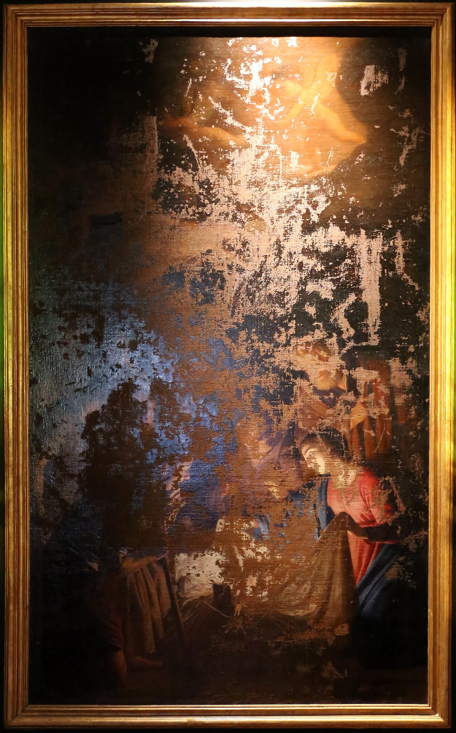 A tall nativity in a gold frame, its paint torn away in large patches. The Virgin in red and blue still kneels on the right, but the centre where the child lay is bare canvas.