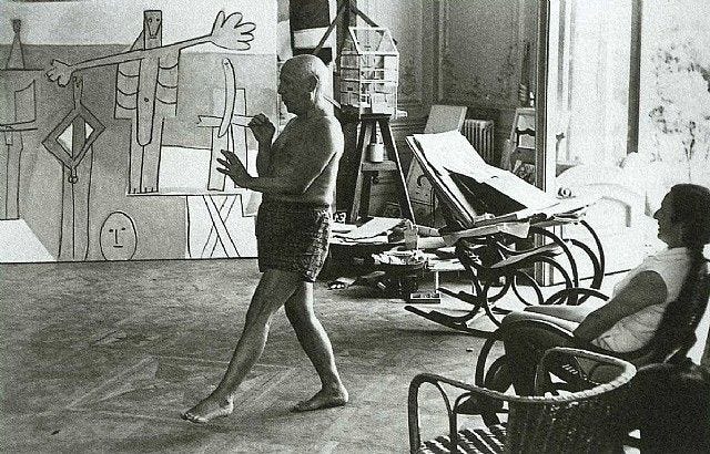 A candid black-and-white photograph of Pablo Picasso in his studio, captured mid-dance, exuding playfulness and creativity. Behind him, large abstract paintings and a cluttered workspace illustrate his prolific artistry, while a seated observer adds a sense of quiet contemplation. This intimate moment showcases the balance between spontaneity and artistic focus.