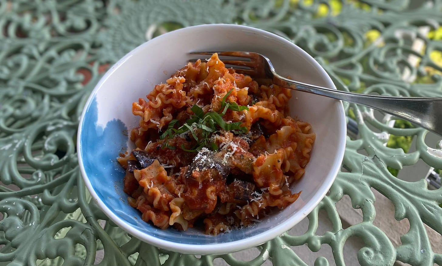 On a mint green outdoor café table, a blue and white bowl of mafalda corta in red sauce with chunks of purple eggplant, topped with grated parmesan and fresh basil. 