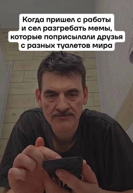 A man with short dark hair and a mustache sits indoors, wearing a black t-shirt, holding a black smartphone close to his face with both hands, looking at the screen intently. The background includes a sink and tiled wall suggesting a bathroom setting. White Russian text overlays the image at the top: Корабль прибыл с тобой и ты распространяешь мемы, which translates contextually to a ship arrived with you and you spread memes. Additional text at the bottom reads: что означает, что ты турист в Урала, meaning that means you are a tourist in the Urals.