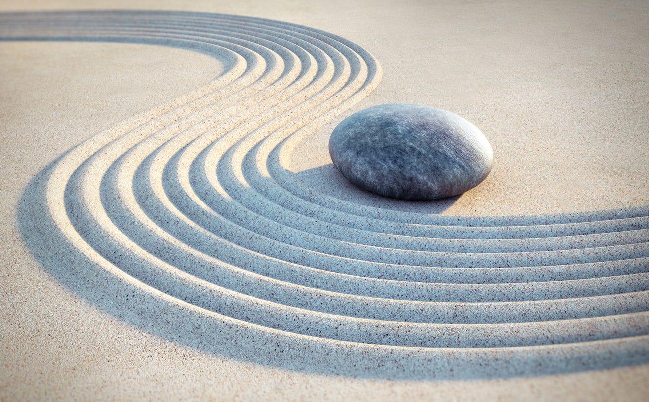 A close up of beige sand with rows of sweeping lines carved in it and one gray, white, and black rock placed within one of its curves. 
