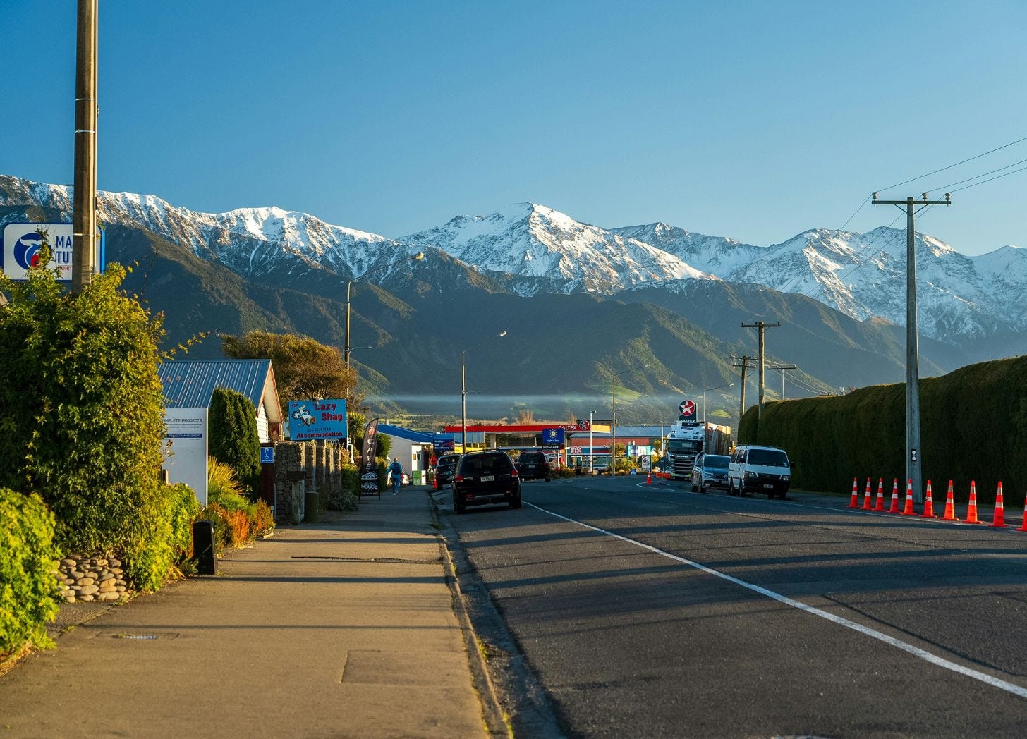 Main street in Kaikōura, New Zealand, with small businesses and snow-capped mountains in the background during a quiet travel period.