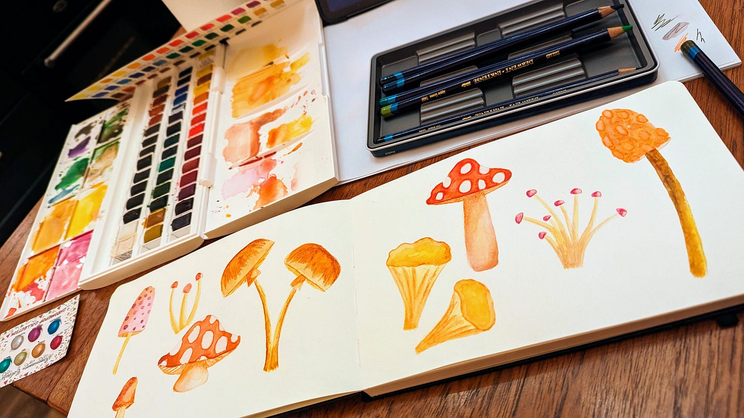 Sketchbook and watercolours on a table with watercolour mushrooms painted