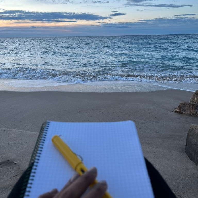 A notebook and a yellow pen beside the ocean as the sun rises, with soft waves in the background — a peaceful setting for writing and reflection.