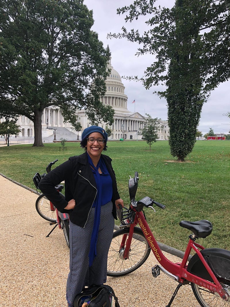 Kristen standing on the East Lawn of the US Capitol, in a blue blouse, a cotton moto jacket and with her hands on her hips, and leaning against a red Captial Bikeshare bicycle. She has on a blue beret and glasses and the US Capitol is in the background on an overcast day. Kristen standing on the East Lawn of the US Capitol, in a blue blouse, a cotton moto jacket and with her hands on her hips, and leaning against a red Captial Bikeshare bicycle. She has on a blue beret and glasses and the US Capitol is in the background on an overcast day.