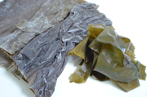 Three types of seaweed displayed side by side: a small bowl of dried mixed seaweed with thin green and dark strands, a thick glossy sheet of kombu, and a stack of crisp square sheets of nori.